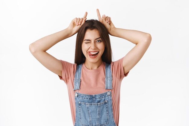 Novia despreocupada, atrevida y descarada que a veces actúa como un demonio mostrando cuernos con los dedos en la parte superior de la cabeza guiñando un ojo descarado y pegando la lengua mientras se pone juguetona sobre fondo blanco