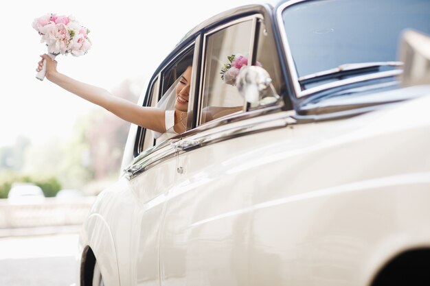 La novia hermosa se sienta con el ramo de la boda en un coche retro y se divierte