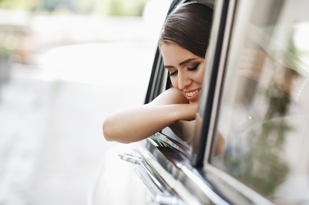 La novia hermosa se sienta con el ramo de la boda en un coche retro y se divierte