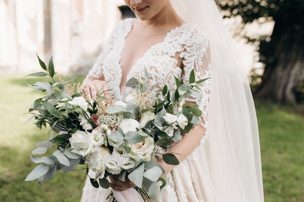 La novia en el hermoso vestido sostiene un ramo de novia con ramas de decoración verde y rosas blancas