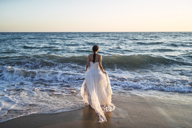 Novia morena caucásica acercándose al mar con un vestido de novia blanco