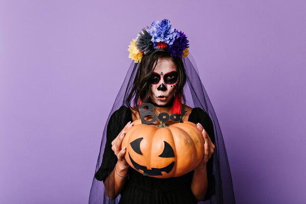 Novia muerta aterradora con calabaza. Mujer europea en velo negro posando en la pared púrpura en halloween.