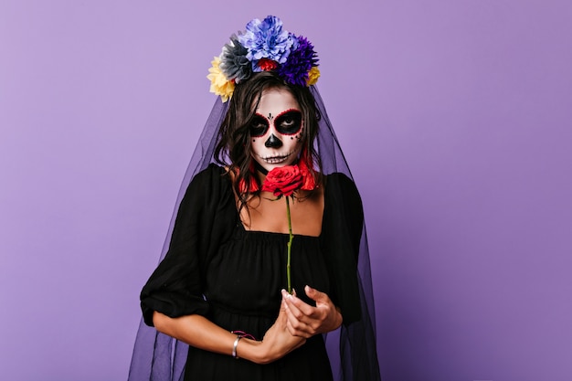 Novia muerta pensativa posando con flores rojas. Filmación en interiores de elegante mujer caucásica en traje de zombie preparándose para la fiesta.