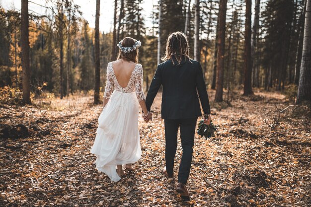 La novia y el novio caminando sobre el suelo cubierto de hojas en el bosque durante el día