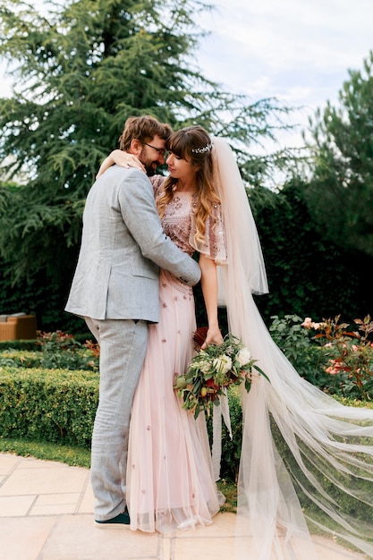 La novia y el novio en el día de la boda caminando al aire libre en la naturaleza de primavera. Feliz recién casada mujer y hombre abrazando en parque verde. Amorosa pareja de novios.