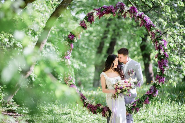 La novia y el novio posan detrás de un gran círculo de lilas en el jardín