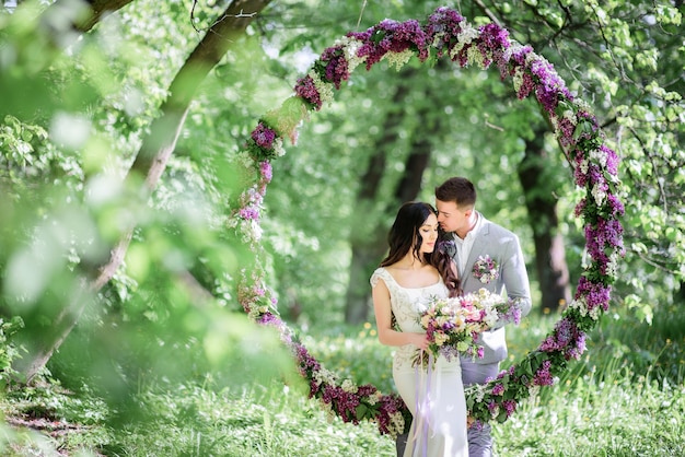 La novia y el novio posan detrás de un gran círculo de lilas en el jardín
