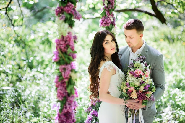 La novia y el novio posan detrás de un gran círculo de lilas en el jardín