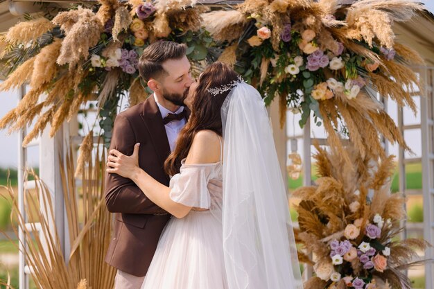 Novia y novio recién casados besándose cerca del arco con hierba pomposa