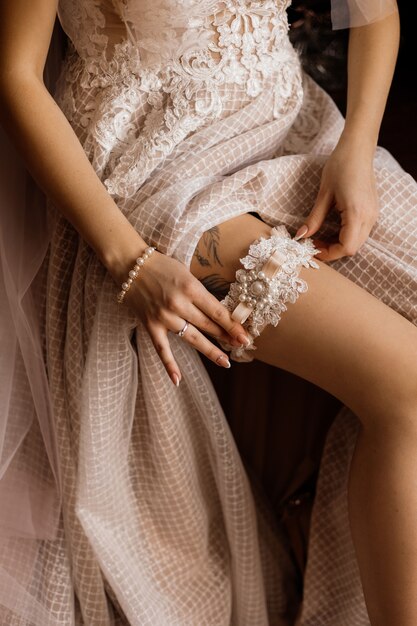 La novia se pone en la pierna la liga de boda tierna, vestida con un vestido de novia con un tatuaje en la pierna