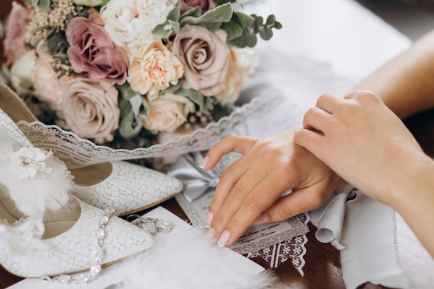 La novia pone sus manos sobre la mesa cerca de un ramo de flores, zapatos y otros detalles de novia.