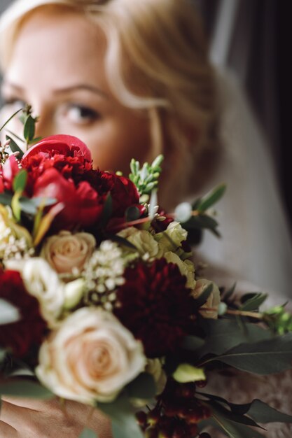 La novia rubia hermosa mira sobre el ramo rojo oscuro de la boda