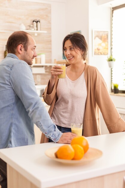 Novia sosteniendo batido nutritivo mientras sonríe al marido en la cocina. Estilo de vida saludable, despreocupado y alegre, comiendo dieta y preparando el desayuno en una acogedora mañana soleada