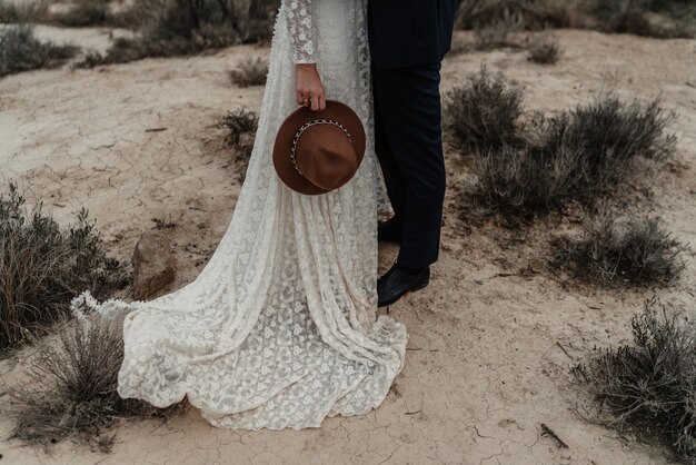 Novia sosteniendo un sombrero y un novio de pie cerca de los arbustos