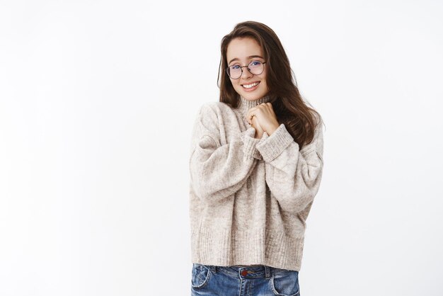 Una novia tonta y encantadora con gafas y suéter cogidas de la mano apretadas contra el pecho y sonriendo con amor sentimientos tiernos estar enamorada de pie tocada y conmovedora mirando con simpatía