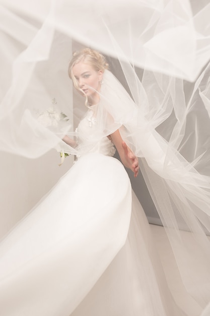 La novia en el vestido hermoso que se coloca dentro en el interior blanco del estudio tiene gusto en casa. tiro de moda estilo boda. joven modelo caucásico atractivo como una novia tierna mirando.