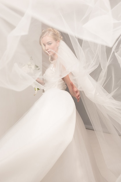 La novia en el vestido hermoso que se coloca dentro en el interior blanco del estudio tiene gusto en casa. Tiro de moda estilo boda. Joven modelo caucásico atractivo como una novia tierna mirando.