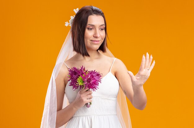 Novia en vestido de novia hermoso con ramo de flores de boda mirando su anillo en su dedo parado sobre pared naranja