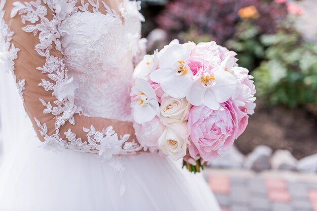 La novia en vestido rico tiene ramo rosado de la boda de orquídeas y peonías