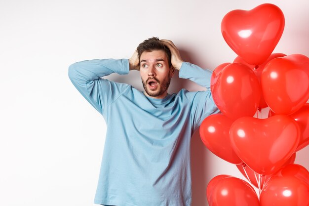 El novio agarra la cabeza entre las manos y entra en pánico por los regalos del día de San Valentín, mirando hacia los lados con cara de alarma, de pie cerca de globos de corazones y pensando en un regalo para el amante, fondo blanco.