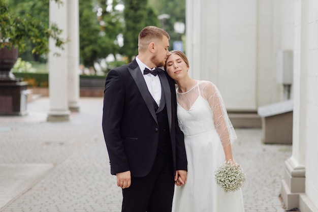 Un novio barbudo y elegante con un traje y una hermosa novia rubia con un vestido blanco con un ramo en sus manos están de pie y abrazándose en la naturaleza en el bosque de pinos.