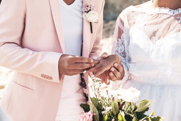 Novio con chaqueta rosa lleva anillo en el dedo de la novia en la playa