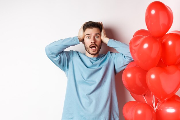 Novio confundido tomados de la mano en la cabeza y en pánico en las vacaciones de San Valentín, alarmado con regalos románticos en el día de los enamorados, de pie cerca del globo de corazones sobre fondo blanco.