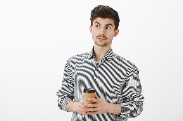 Novio europeo maduro confundido con bigote y barba, mirando hacia otro lado con las cejas levantadas, tomando café y siendo interrogado con el comportamiento de un amigo, pensando que es extraño sobre una pared gris