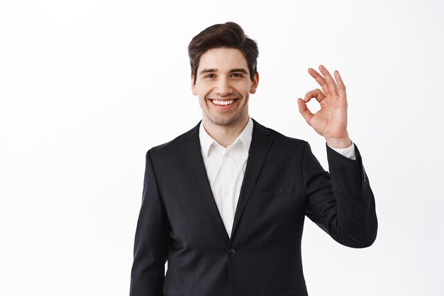 Novio guapo mostrando signo bien. El hombre de negocios en traje negro hace el gesto de OK y sonriendo satisfecho, elogia el buen trabajo, bien hecho, excelente gesto, fondo blanco.