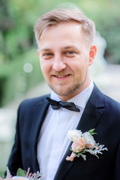 Novio guapo en traje negro y boutonniere rosa blanca se encuentra en el jardín