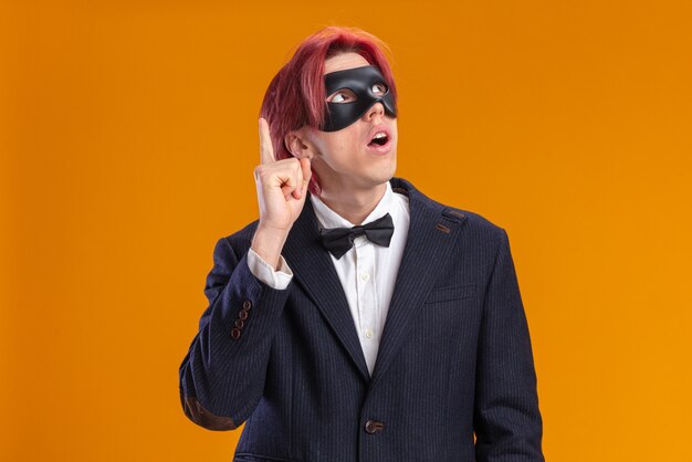 Novio guapo en traje con pajarita y máscara de mascarada mirando sorprendido apuntando con los dedos índices hacia arriba