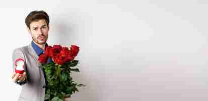 Foto gratuita novio guapo en traje pidiendo casarse con él de pie con ramo rojo de rosas y anillo de compromiso