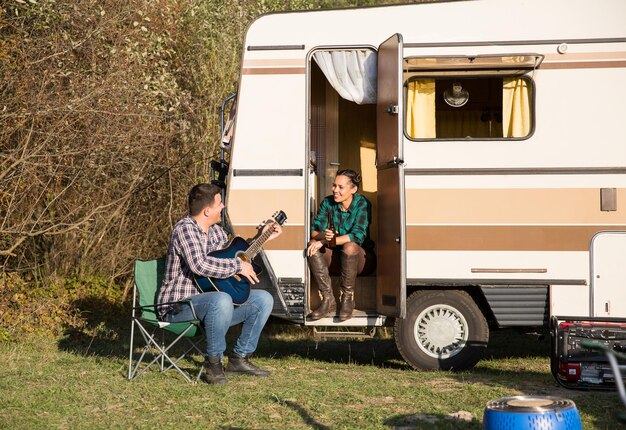 Novio hipster tocando una vieja guitarra a su novia frente a su autocaravana retro en las montañas.