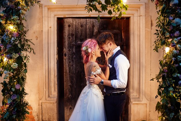 Novio y novia con cabello rosado parado frente a una puerta con guirnaldas de flores y luz