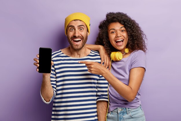 El novio y la novia emocionales diversos muestran un moderno dispositivo de teléfono inteligente con una pantalla simulada para su contenido promocional