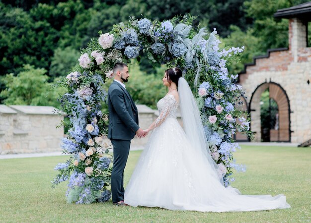 El novio y la novia están de pie juntos frente al arco decorado con hortensias azules, tomados de la mano, ceremonia de boda, votos matrimoniales