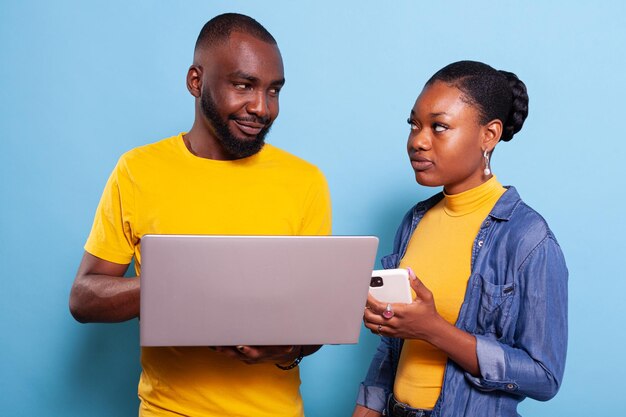 Novio y novia trabajando con computadora y teléfono móvil en estudio. Personas en relación que usan laptop y smartphone con tecnología moderna, mirándose en la cámara.