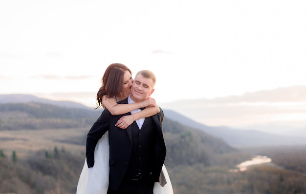 El novio sonreído está sosteniendo a la novia en la espalda al aire libre con colinas en el fondo al atardecer