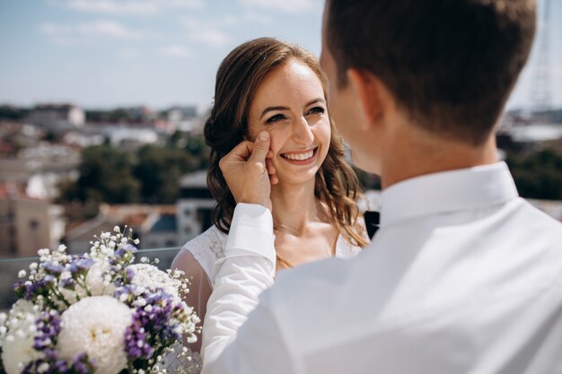 El novio sostiene a la novia de pie en el techo con un gran paisaje urbano de verano a su alrededor