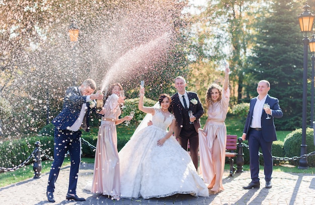 Novios y mejores amigos beben champaña y celebran en el parque el día de la boda