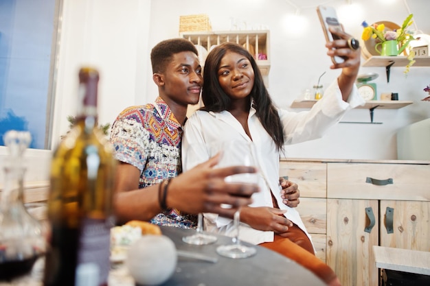 Novios de pareja afroamericana bebiendo vino en la cocina en su cita romántica con el teléfono móvil en las manos