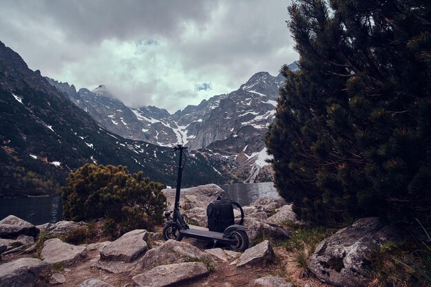 Nubes bajas oscuras, hermosas montañas, lago, pinos y mochila con scooter en el frente.