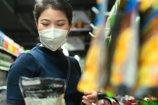 Nueva normalidad después de la epidemia de covid, joven mujer asiática inteligente que compra un nuevo estilo de vida en el supermercado con protector facial o máscara de protección, elija productos de consumo, nuevo estilo de vida normal