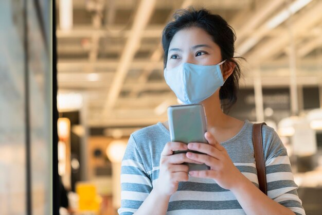 Nueva normalidad después de la epidemia de covid19, máscara de protección de uso femenino asiático inteligente, uso manual de la comunicación del teléfono inteligente en los grandes almacenes, nuevo estilo de vida, fondo borroso abstracto