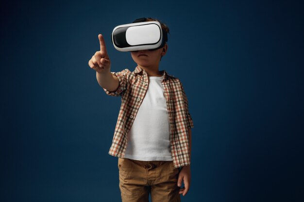 Nuevas ideas y emociones. Niño o niño apuntando al espacio vacío con gafas de realidad virtual aisladas sobre fondo blanco de estudio. Concepto de tecnología de punta, videojuegos, innovación.
