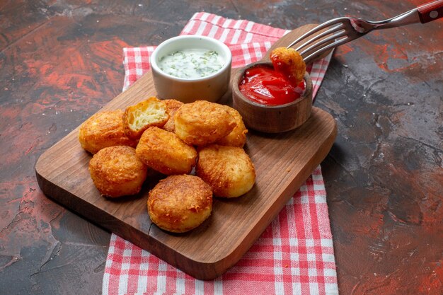 Nuggets de pollo de vista inferior sobre tablero de madera con pepitas de salsas en horquilla sobre mesa oscura