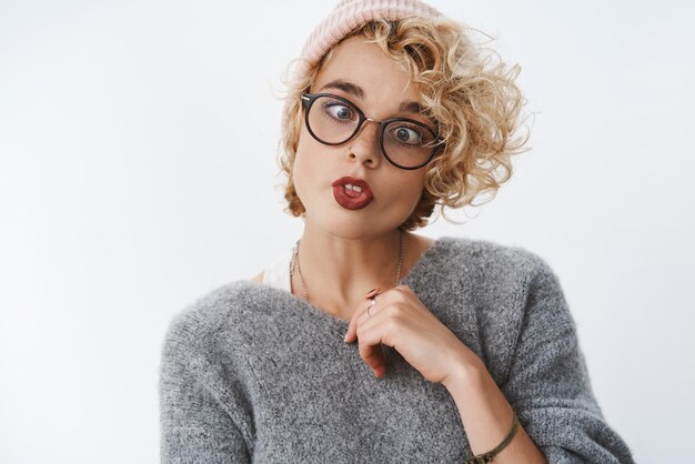 Nunca es demasiado mayor para perder el tiempo. Retrato de novia rubia linda divertida juguetona e inmadura en gorro y suéter de invierno entrecerrando los labios plegables y mostrando la lengua en la pared blanca. Copia espacio