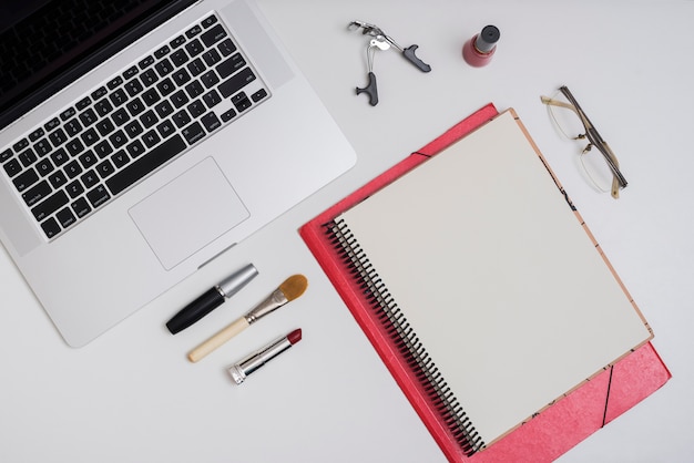 Objetos de maquillaje con carpetas portátiles y de oficina; lente en el escritorio blanco