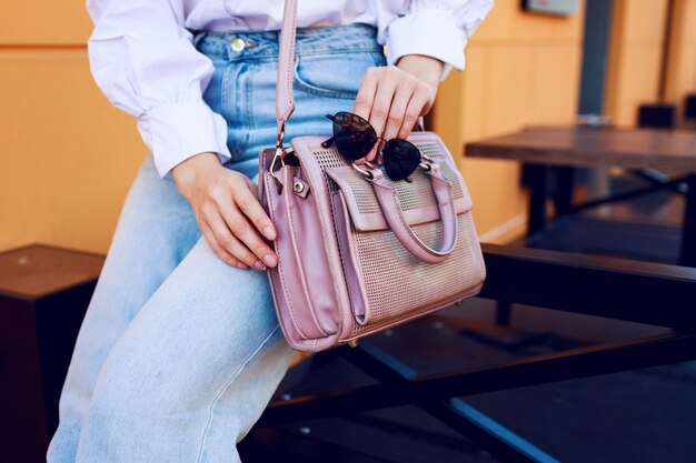 Objetos de moda. Manos de mujer con bolso y gafas de sol. Chica de moda sentado al aire libre. Jeans con estilo.