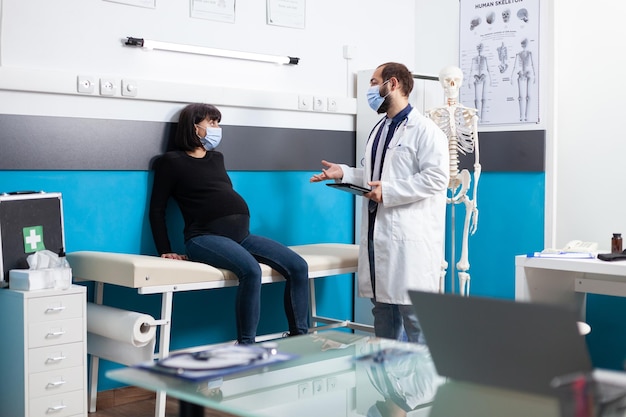 Obstetra con dispositivo que brinda apoyo médico a pacientes embarazadas, haciendo consultas de atención médica en el consultorio. Mujer embarazada que recibe apoyo de maternidad de un médico con mascarilla.